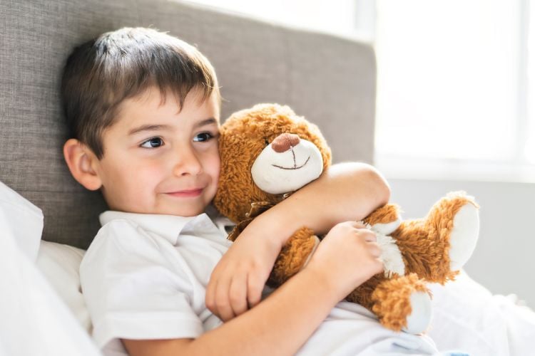 Um menino está deitado em uma cama, abraçando um ursinho de pelúcia. Ele exibe um sorriso suave, transmitindo conforto e segurança. O ambiente é iluminado e acolhedor. 