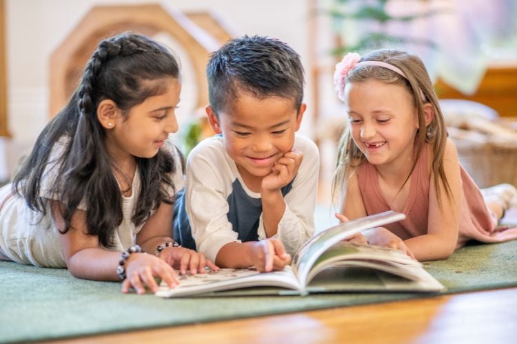 Três crianças estão deitadas no chão, sorrindo enquanto folheiam um livro. O ambiente é acolhedor, com luz suave e decoração infantil. 