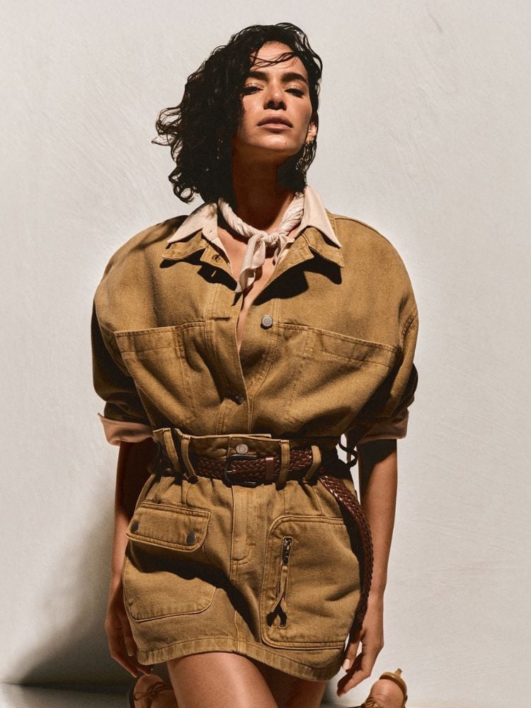 Bruna Marquezine posando em um cenário de luz suave e sombra. Ela está vestindo uma roupa utilitária de estilo militar, feita de tecido em tom marrom-terroso. A peça parece ser uma jaqueta oversized com bolsos grandes e um zíper frontal, usada como um vestido curto, combinada com um cinto de couro trançado marrom que define sua cintura. O cabelo dela é curto e ondulado, e ela usa uma bandana clara ao redor do pescoço.