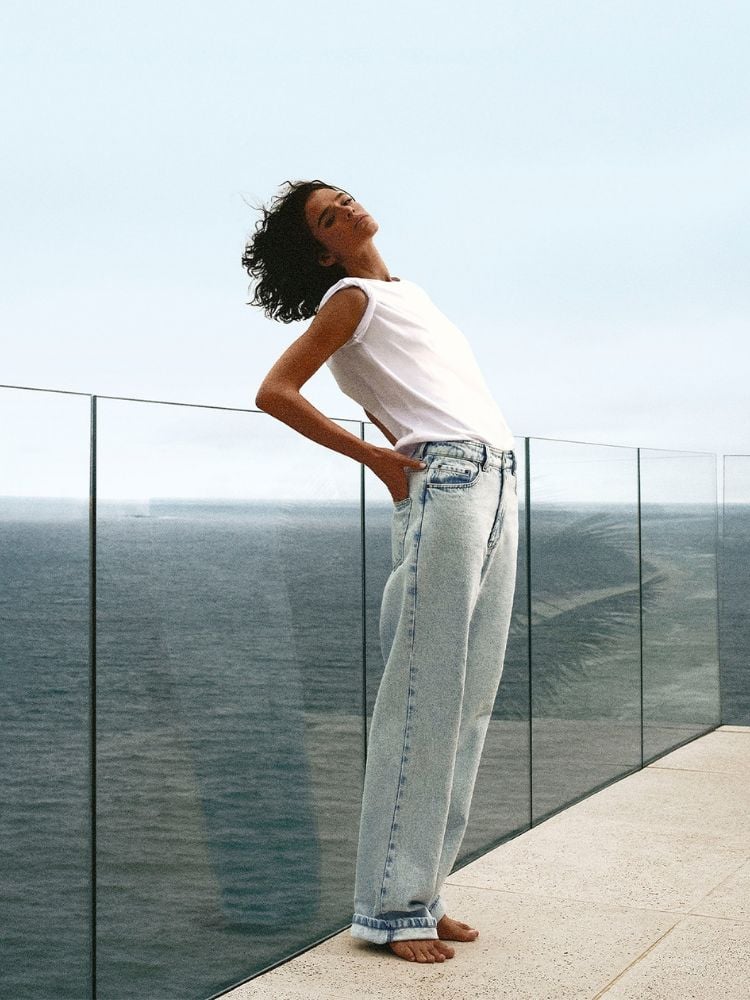 A imagem mostra a atriz Bruna Marquezine em uma pose descontraída, inclinando o corpo para trás. Ela está em uma varanda com piso claro e um parapeito de vidro, com vista para o mar no Rio de Janeiro. Bruna usa um look casual composto por uma camiseta branca básica e uma calça jeans de cintura alta e corte reto, de tonalidade clara e desgastada e ela está descalça.