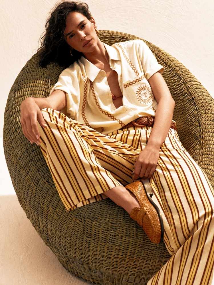 Na imagem, Bruna Marquezine está sentada de maneira relaxada em uma poltrona de palha arredondada, com um estilo casual e sofisticado. Ela veste uma camisa de botões em tons claros com detalhes em dourado, incluindo um desenho de sol. A camisa está parcialmente desabotoada. Bruna também usa uma calça ampla listrada em tons de amarelo e marrom, com o cinto de couro marrom que marca sua cintura. Nos pés, ela calça sapatos estilo mocassim em tom marrom trançado. 