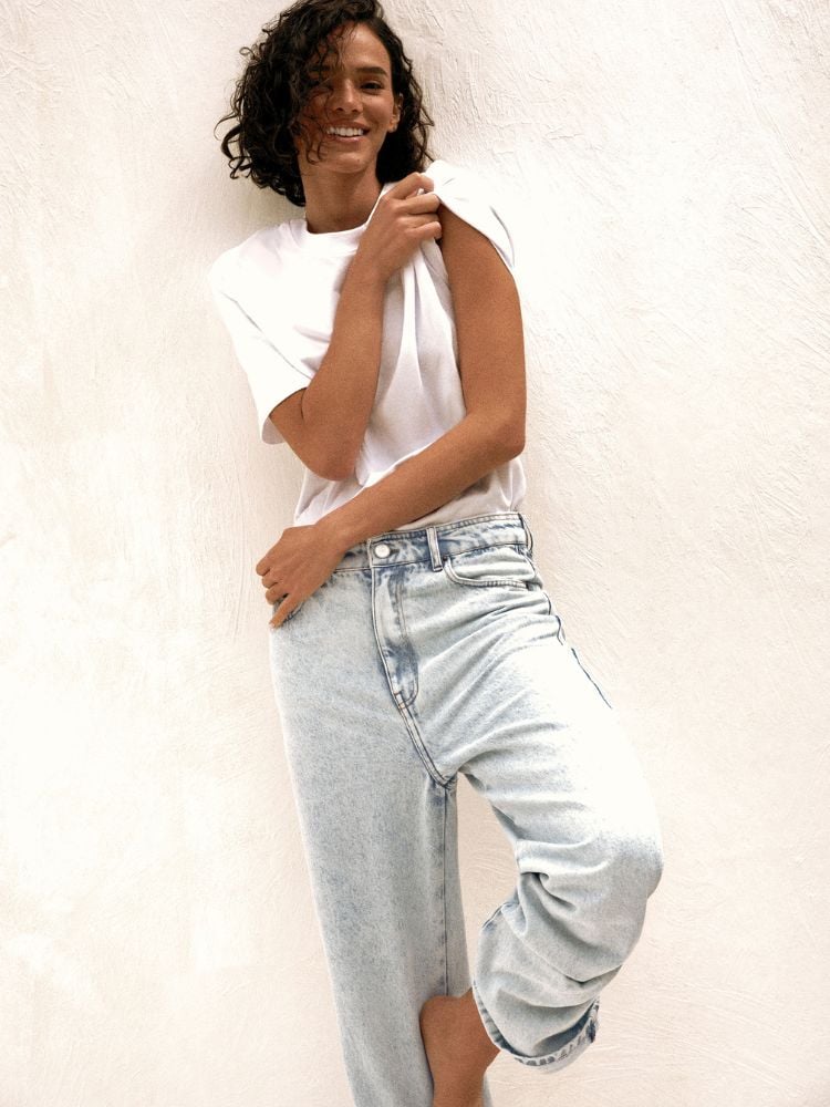 A imagem mostra Bruna Marquezine em uma pose casual e descontraída. Ela veste uma camiseta branca básica, ligeiramente folgada, combinada com uma calça jeans de lavagem clara, com corte também folgado e barra dobrada, criando um look despojado e confortável. Bruna sorri de maneira espontânea e está com uma postura relaxada, segurando a manga da camiseta com uma das mãos, enquanto cruza uma perna. O fundo é neutro, em um tom claro, destacando o estilo minimalista da composição.