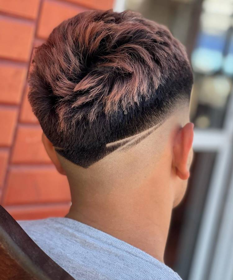 O corte de cabelo masculino apresenta um estilo moderno com finalização em V na nuca. O cabelo no topo é volumoso e texturizado, com coloração em tons rosados. As laterais exibem um fade gradual, destacando uma linha geométrica raspada que adiciona um toque de criatividade. O visual é contemporâneo e arrojado, ideal para quem busca um estilo ousado.