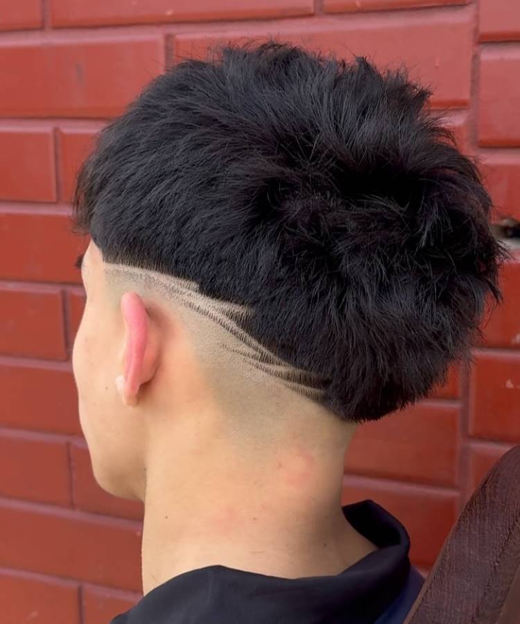 O corte de cabelo masculino apresenta um estilo moderno com um topo volumoso e texturizado. As laterais exibem um fade acentuado, destacando linhas geométricas raspadas que criam um design dinâmico. A parte inferior é mais curta, realçando o contraste entre as diferentes seções. O visual é arrojado e contemporâneo, ideal para quem busca um estilo único e criativo.