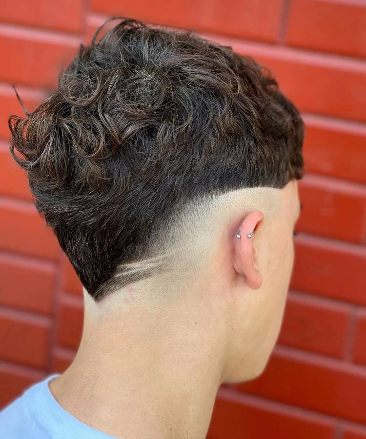 O corte de cabelo masculino apresenta um topo cacheado e volumoso, com um fade acentuado nas laterais. A nuca exibe um design em V, complementado por uma linha geométrica raspada. O visual é moderno e estiloso, destacando-se pelo contraste entre os cabelos mais longos no topo e o acabamento preciso nas laterais e na parte inferior.