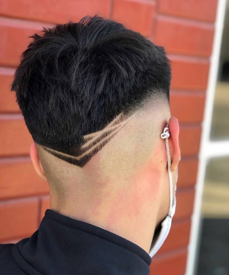 O corte de cabelo masculino exibe um topo volumoso com um fade acentuado nas laterais e na nuca. A parte inferior apresenta um design em V, complementado por linhas geométricas raspadas que criam um efeito dinâmico e moderno. O visual é arrojado e estiloso, ideal para quem busca um toque de criatividade no penteado.