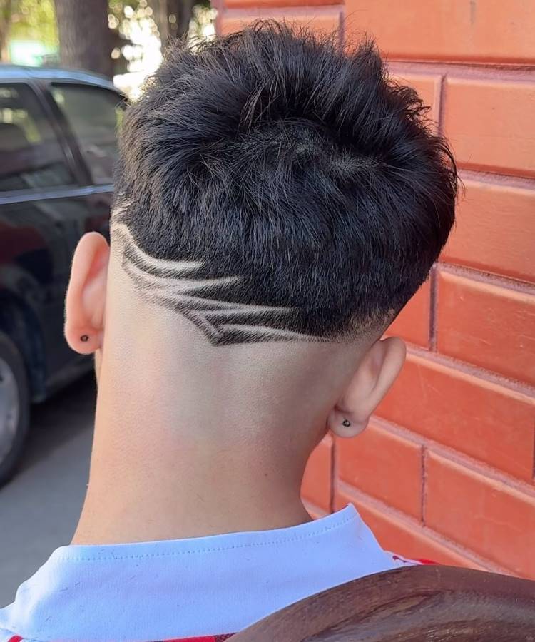 O corte de cabelo masculino apresenta um topo volumoso e texturizado. As laterais e a nuca têm um fade acentuado, destacando um design geométrico raspado. O desenho cria linhas dinâmicas que adicionam um toque moderno e estiloso ao visual. A combinação do volume superior com o detalhe artístico na nuca torna o estilo ousado e contemporâneo.