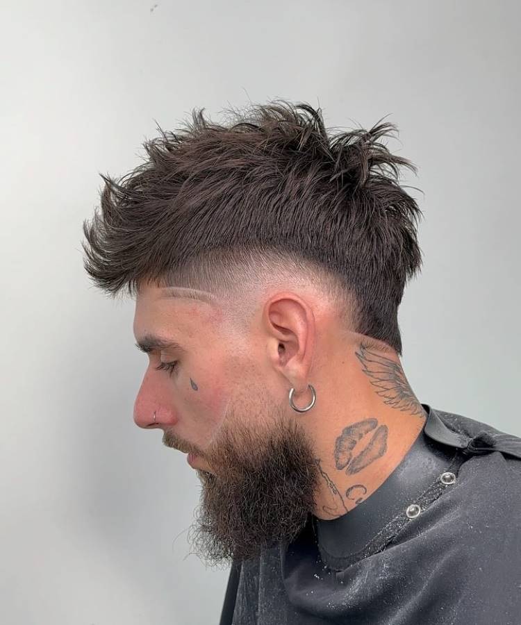 O corte de cabelo masculino apresenta um topo volumoso e texturizado, com um fade acentuado nas laterais. Uma linha raspada adiciona um detalhe geométrico moderno. O visual é complementado por uma barba cheia e bem cuidada. O homem tem tatuagens visíveis no pescoço, incluindo asas e lábios, além de um brinco grande na orelha esquerda.