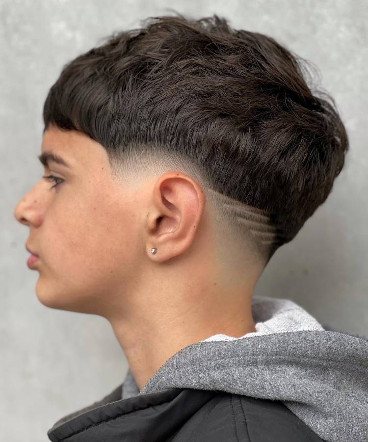 O jovem tem um corte de cabelo moderno com volume no topo e um fade acentuado nas laterais. O cabelo é escuro e liso, com uma linha geométrica raspada na nuca, adicionando um toque estiloso. Ele usa um brinco discreto na orelha esquerda e veste uma jaqueta preta sobre um moletom cinza, criando um visual casual e contemporâneo.