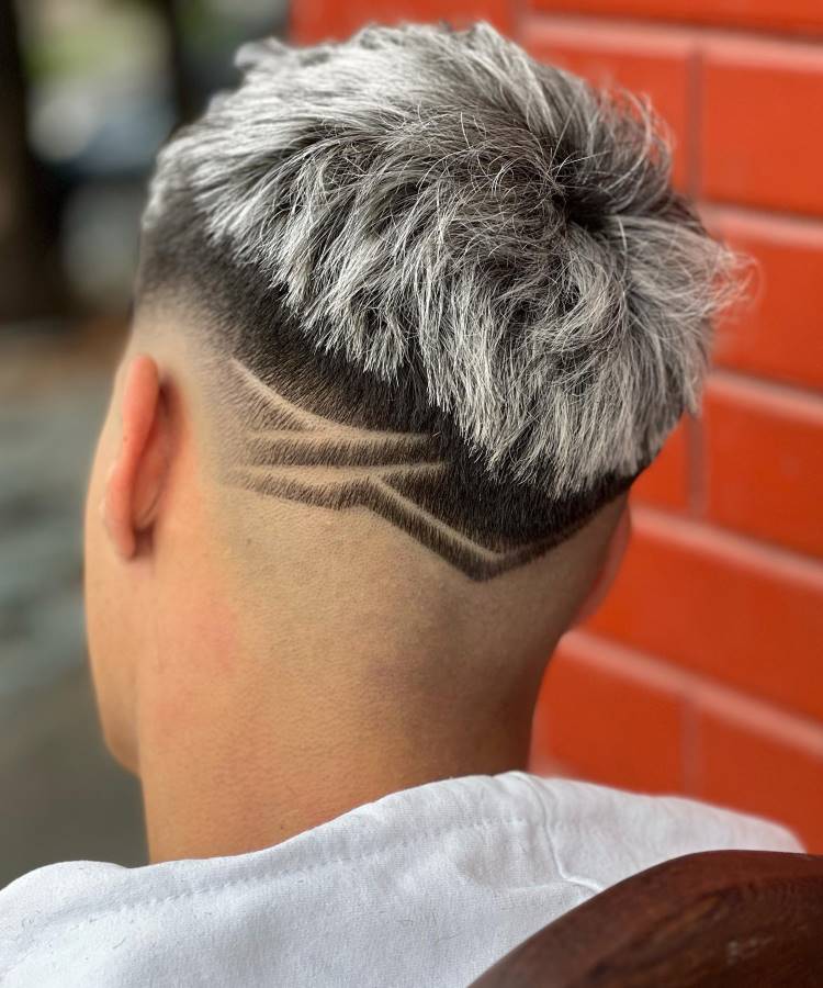 O corte de cabelo masculino exibe um topo volumoso e platinado, com um fade acentuado nas laterais. A nuca é decorada com linhas geométricas raspadas, formando um design dinâmico e moderno. O contraste entre o cabelo claro e os detalhes escuros cria um visual arrojado. O fundo da imagem mostra uma parede de tijolos vermelhos, adicionando cor ao cenário.