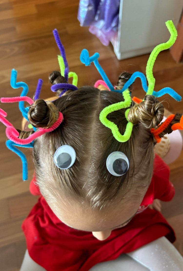 Uma criança está com um penteado divertido, usando limpadores de cachimbo coloridos enrolados no cabelo, criando um efeito de antenas. Dois olhos de brinquedo estão colados na testa, dando um visual de criatura. O cabelo está dividido ao meio e preso em coques. O ambiente tem piso de madeira e a criança veste vermelho.