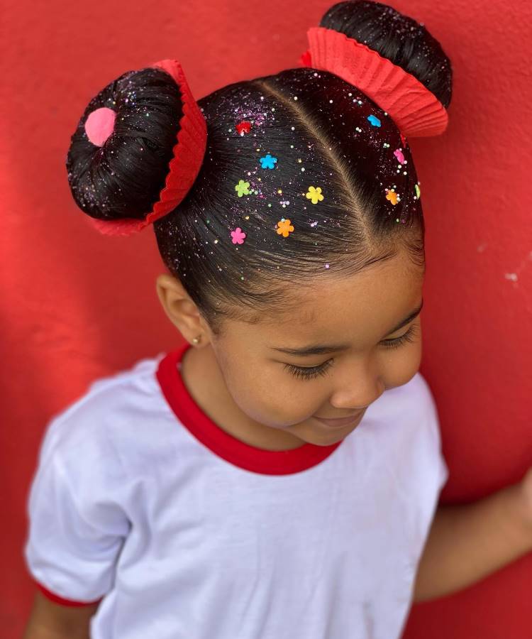 A imagem mostra uma menina com um cabelo maluco, estilizado em dois coques decorados com forminhas de cupcake vermelhas. Os coques têm detalhes coloridos, como confeitos e glitter, que dão um toque divertido e criativo ao penteado. O fundo vermelho e a camiseta branca com detalhes vermelhos complementam o visual alegre e vibrante.