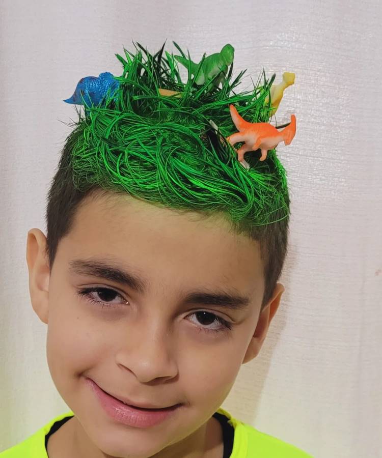 A imagem mostra um menino com cabelo maluco, pintado de verde vibrante e estilizado para parecer grama. No topo, há pequenos dinossauros coloridos, criando um cenário divertido e criativo. O penteado evoca uma sensação de aventura e imaginação, destacando-se contra a camiseta amarela brilhante que ele está vestindo.