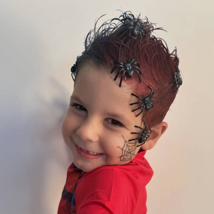 A imagem mostra uma criança sorridente com um cabelo maluco, tingido de vermelho e decorado com aranhas de brinquedo. O penteado é ousado e divertido, com as aranhas estrategicamente posicionadas para criar um visual de teia. Na bochecha, há uma pintura de teia de aranha, complementando o tema assustador e criativo do look.