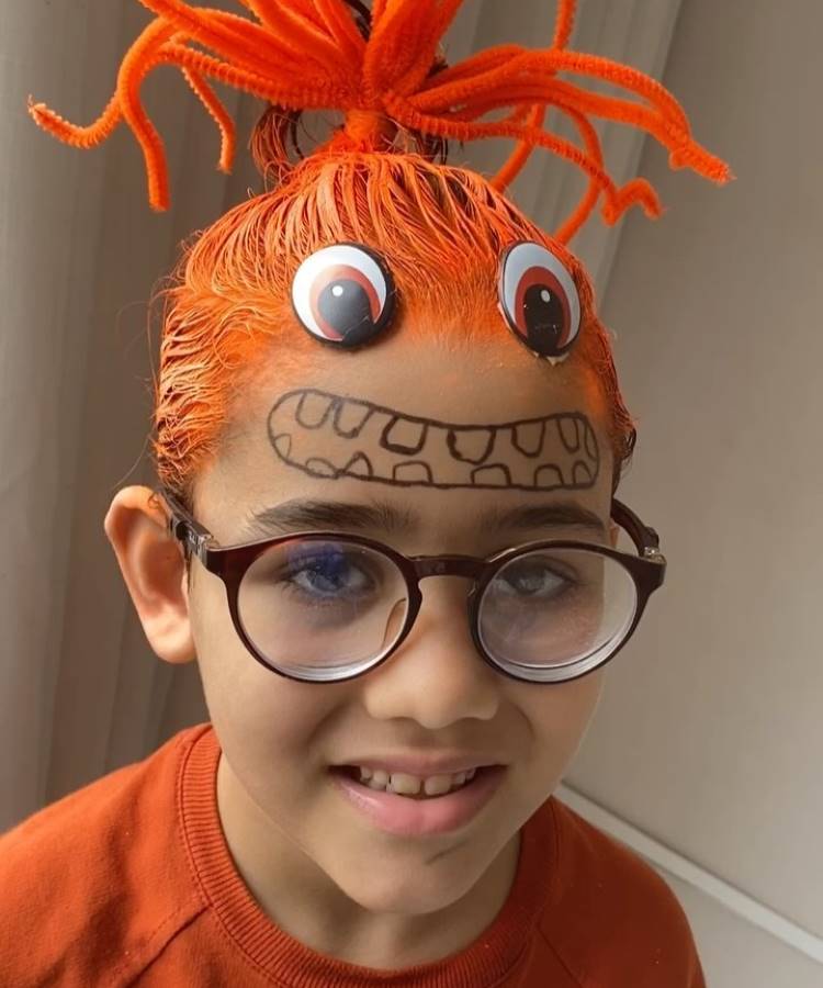 A imagem mostra uma criança sorridente com um penteado criativo e divertido. O cabelo é tingido de laranja vibrante, preso no alto com fios formando um coque espetado. Dois grandes olhos de brinquedo estão fixados na testa, e uma boca desenhada completa o visual de monstro. A criança usa óculos e veste uma camiseta laranja.