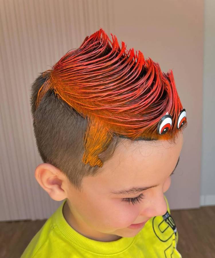 A imagem mostra um menino com cabelo maluco, estilizado em um moicano vermelho e laranja vibrante. O cabelo é penteado em pontas levantadas, criando um visual ousado e divertido. Dois olhos esbugalhados estão presos na parte da frente, dando um efeito de monstro. O menino veste uma camiseta amarela e sorri levemente.