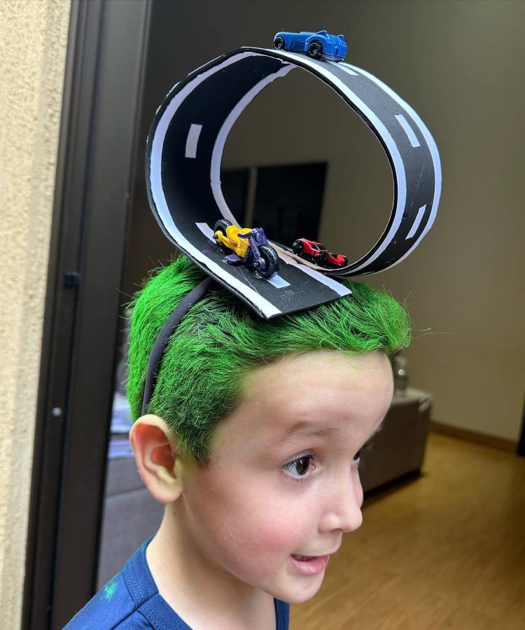 A imagem mostra um menino com cabelo maluco, pintado de verde vibrante. No topo da cabeça, há uma pista de corrida em miniatura, completa com carros de brinquedo e uma moto. A pista forma um loop impressionante, criando um visual divertido e criativo. O menino está sorrindo, mostrando entusiasmo com seu penteado único.