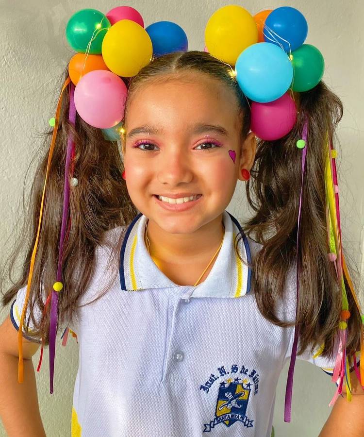 A imagem mostra uma menina sorridente com um penteado criativo. Seu cabelo está preso em duas maria-chiquinhas decoradas com balões coloridos, incluindo amarelo, azul, verde e rosa. Fitas coloridas pendem dos cabelos, complementando o visual. Ela usa uma camisa polo branca com detalhes em amarelo e azul, e um pequeno coração está desenhado em seu rosto.