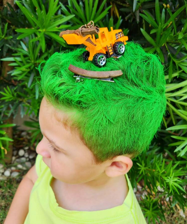 A imagem mostra uma criança com cabelo maluco, tingido de verde brilhante. No topo, há um brinquedo de caminhão de construção amarelo, criando um efeito divertido. A criança veste uma camiseta amarela e está ao ar livre, cercada por folhagens. O visual é criativo e chama a atenção pelo contraste entre o cabelo colorido e os acessórios.