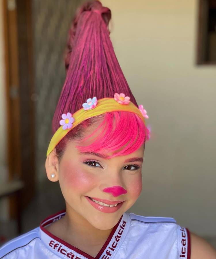 A imagem mostra uma menina com cabelo maluco, estilizado em um cone alto e rosa vibrante. Ela usa uma faixa amarela com flores coloridas, complementando o visual divertido. O rosto está maquiado com blush rosa e um nariz pintado da mesma cor. Ela veste uma camiseta branca e sorri para a câmera.