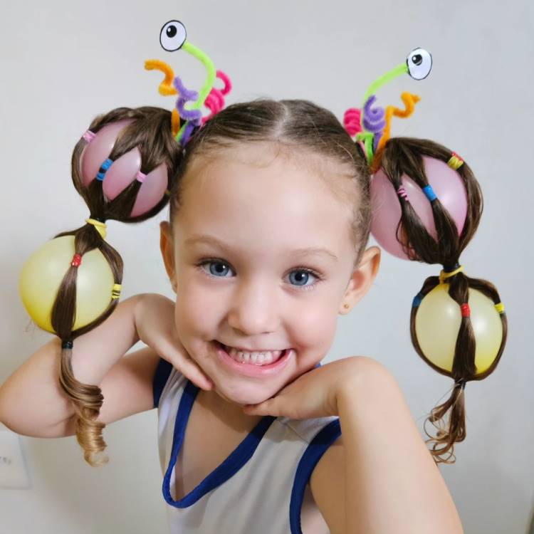 A imagem mostra uma menina com cabelo maluco, estilizado com balões coloridos presos em tranças. Os balões formam grandes pompons em cada lado da cabeça. Adornos de olhos esbugalhados e limpadores de cachimbo coloridos completam o visual criativo e divertido. A menina sorri, apoiando o queixo nas mãos, transmitindo alegria e descontração.