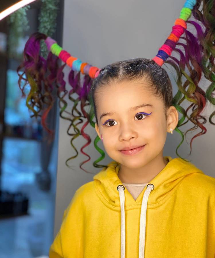 A imagem mostra uma criança com um penteado vibrante e criativo. O cabelo está dividido em dois coques elevados, decorados com fitas coloridas em tons de roxo, verde, vermelho e azul. As pontas são encaracoladas. A criança veste um moletom amarelo e tem um sorriso suave, com maquiagem discreta nos olhos. O fundo é desfocado, destacando o visual.