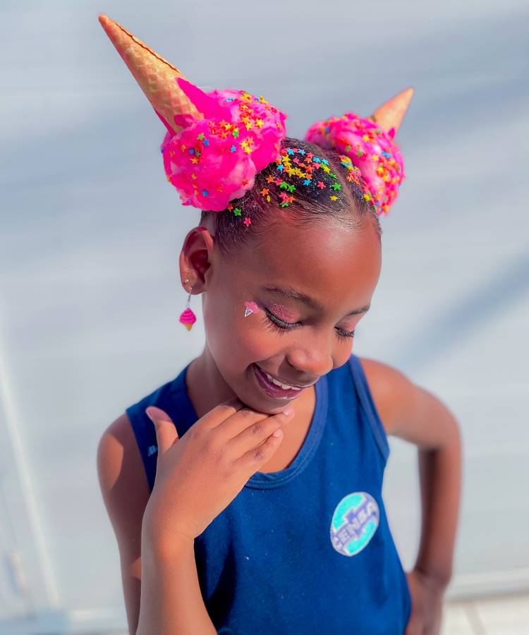 A imagem mostra uma menina sorridente com um penteado criativo, simulando sorvetes. Dois coques grandes e rosa são decorados com granulados coloridos e cones de sorvete. Ela usa uma blusa azul e brincos pendentes. A expressão dela é alegre, e o visual vibrante destaca-se pelo uso de cores vivas e acessórios divertidos.