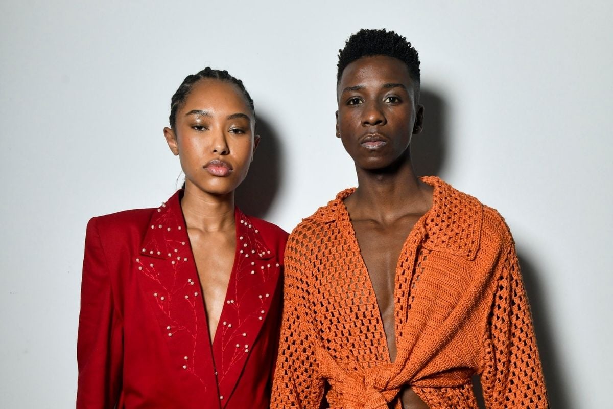 Foto de duas modelos em frente a parede branca, uma usando blazer vermelho e outra usando camisa de crochê terracota