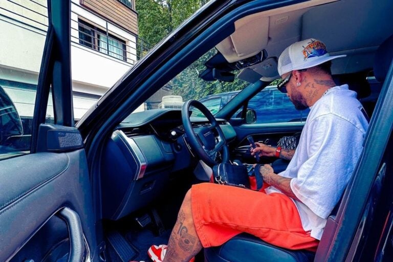 Foto de Neymar Jr dentro de Range Rover Fisrt Edition preta, usando boné branco, blusa branca e bermuda laranja