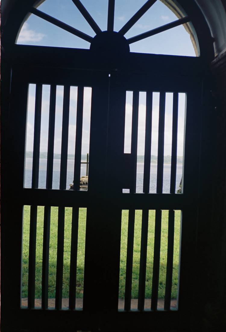 A imagem mostra a vista através de uma porta de madeira antiga, com barras verticais e um arco superior. Além da porta, um campo verde leva ao horizonte, onde um rio ou mar é visível sob um céu azul claro. O cenário evoca a tranquilidade e a história das casas centenárias da ilha.