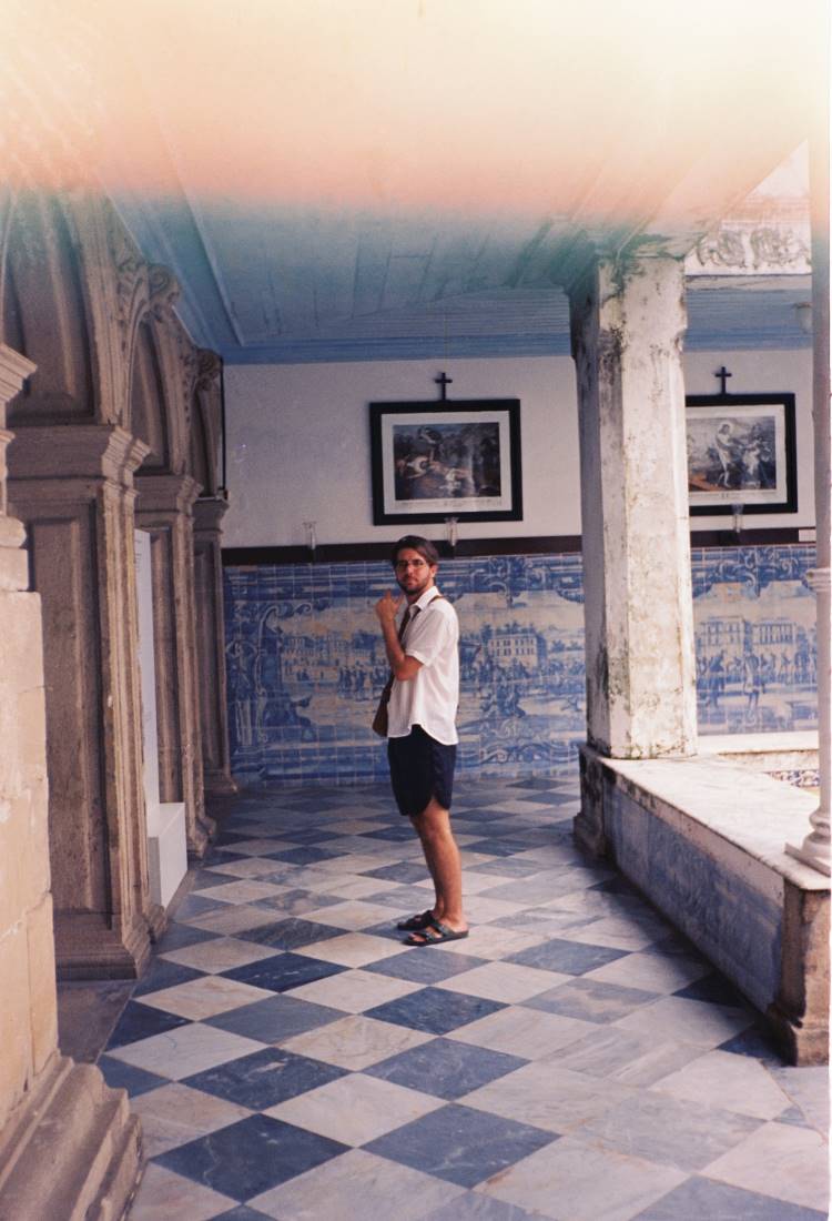 A imagem mostra um homem em um corredor de uma casa centenária, com piso de azulejos em padrão xadrez azul e branco. As paredes são adornadas com azulejos decorativos e quadros. Colunas antigas sustentam o teto, criando um ambiente histórico e artístico, típico das construções tradicionais da ilha.