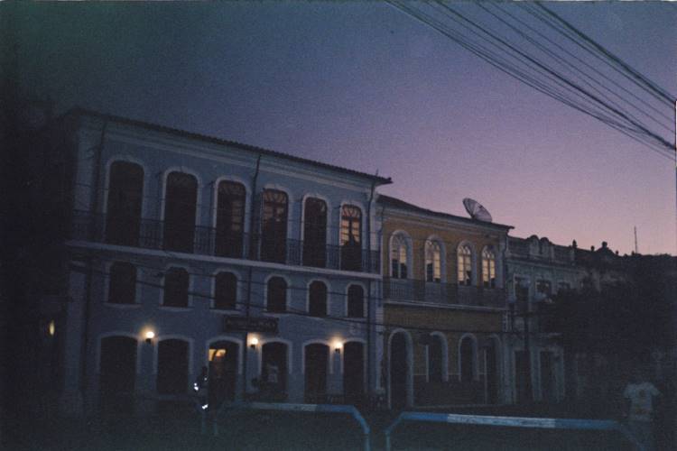 A imagem captura casas centenárias de uma ilha ao entardecer, com fachadas elegantes e janelas arqueadas. As construções de dois andares exibem detalhes arquitetônicos clássicos, enquanto a iluminação suave destaca suas formas. O céu em tons de azul e roxo cria um ambiente nostálgico, evocando a história e o charme das residências antigas.