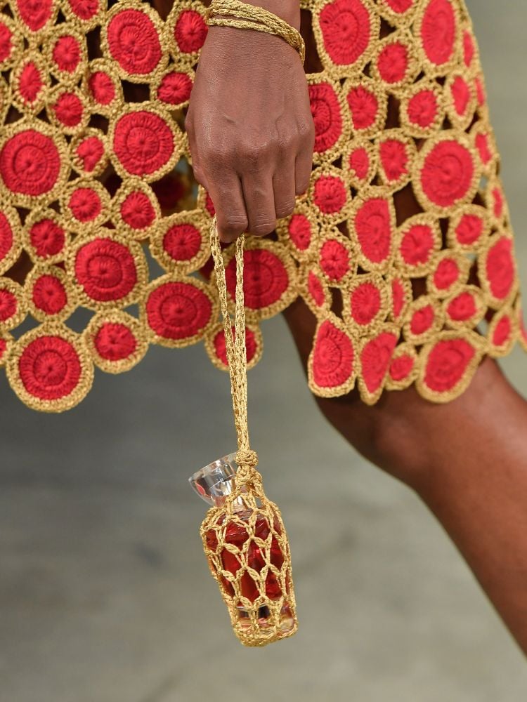 Zoom em bolsa vermelha e dourada, carregada por pessoa de pele negra que usa vestido vermelho e dourado