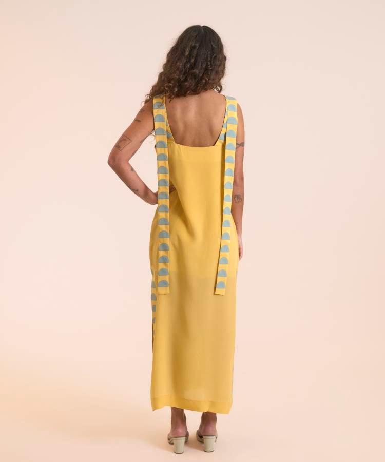 A imagem apresenta uma modelo de costas usando um vestido longo amarelo da coleção Paraguaçu da Handred. O vestido possui alças largas com estampas geométricas em azul claro. A peça destaca um design minimalista e elegante, refletindo a inspiração no Rio Paraguaçu. A modelo está em um fundo neutro, acentuando a simplicidade e sofisticação do look.