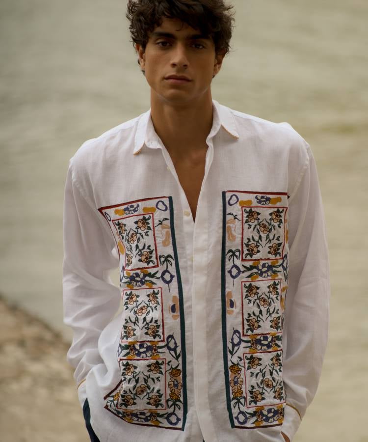 A imagem mostra um modelo vestindo uma camisa branca da coleção Paraguaçu da Handred. A peça é adornada com bordados florais coloridos em tons de azul, amarelo e verde, criando um contraste vibrante. O tecido leve e a abertura frontal refletem um estilo casual e sofisticado, inspirado na beleza natural e cultural do Rio Paraguaçu.