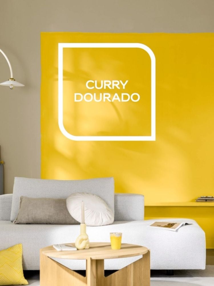 Foto de sala com parede cinza e amarela "curry dourado" da Coral, além de sofá off white com almofadas e mesa de centro cinza com itens decorativos amarelos 