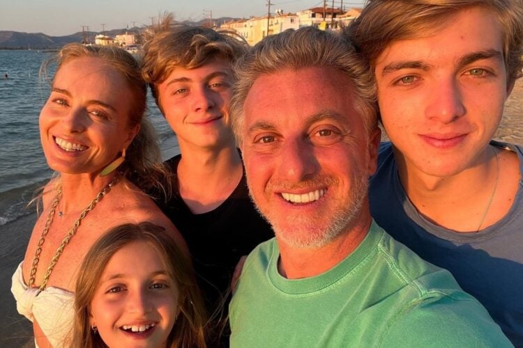 Na foto, há cinco pessoas sorrindo em uma praia ao pôr do sol. O mar está ao fundo, e todos parecem felizes e relaxados. Uma mulher loira está à esquerda, usando um colar dourado. Ao lado dela, um jovem de cabelo loiro e ondulado. No centro, um homem de cabelo grisalho e barba, vestindo uma camiseta verde. À direita, outro jovem, também loiro. Na frente, uma menina loira com um sorriso largo. O cenário é iluminado pela luz dourada do sol.