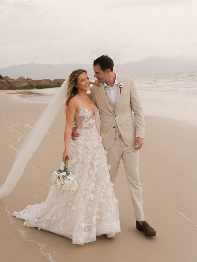 Mulher de pele clara usando véu, vestido de noiva de tule com bordados 3D e homem de terno bege, comemorando casamento na areia da praia  