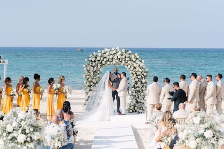 Casamento na praia com arco de flores, noivos, madrinhas de amarelo, padrinhos e convidados sentados 