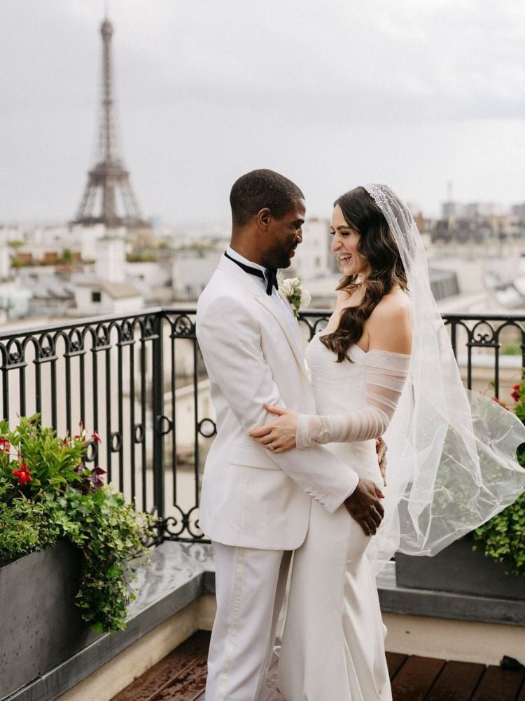 Homem de pele negra usando terno branco e mulher de descendência árabe de noiva, casando-se com a Torre Eiffel ao fundo 