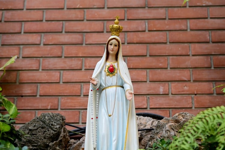 Estátua de Nossa Senhora das Graças com roupa branca e muro de tijolos à vista atrás