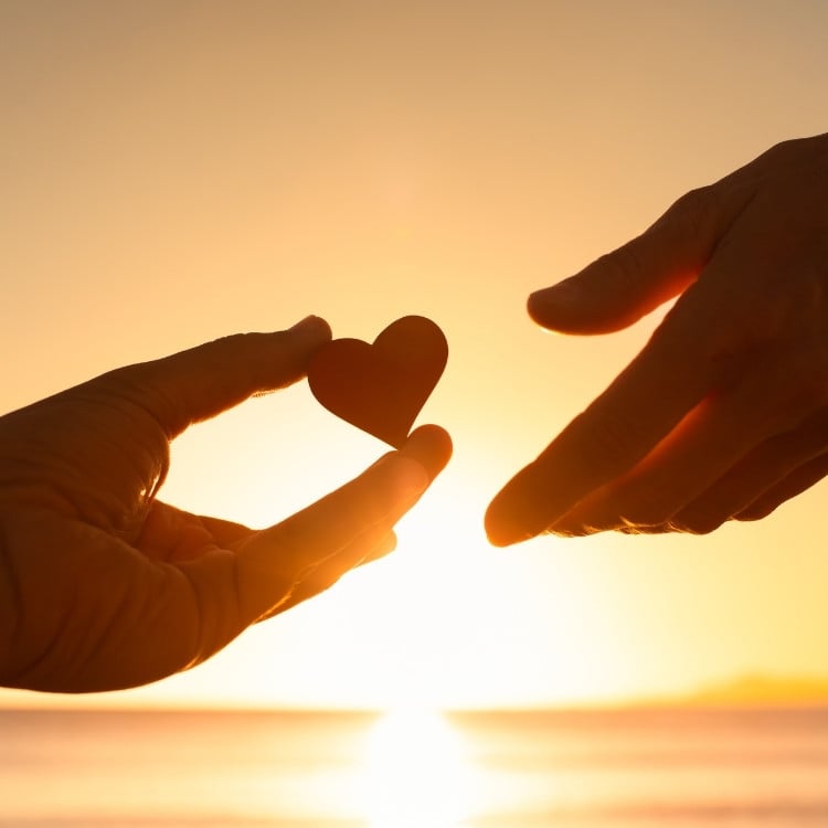 Foto de duas mãos, sendo uma dela segurando um coração de papel, em pôr do sol 