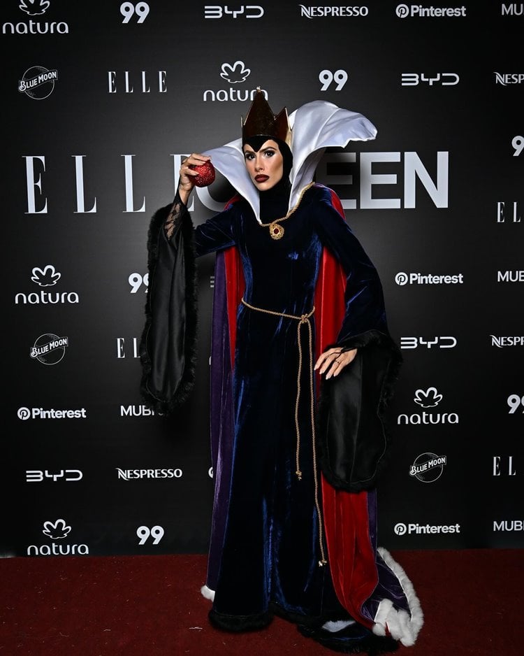 Dy Miryan está vestida como a Rainha Má de "Branca de Neve" para o Elleoween. Ela usa um manto longo roxo com forro vermelho e uma gola alta branca. A coroa dourada e a maçã vermelha em sua mão são detalhes icônicos. A maquiagem é marcante, com olhos delineados e lábios vermelhos.