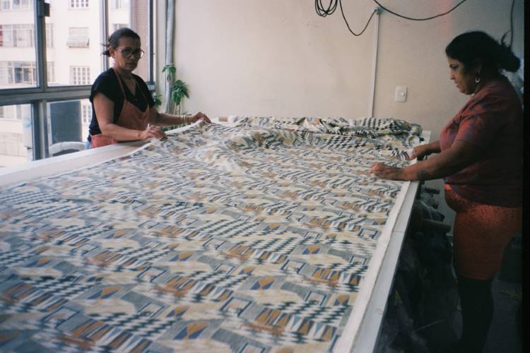 Duas mulheres trabalham em um grande tecido estampado, ajustando-o cuidadosamente sobre uma mesa. A estampa geométrica é rica em detalhes, refletindo o processo artesanal da nova coleção da Handred. A luz natural entra pela janela, iluminando o ambiente de trabalho e destacando a dedicação e o cuidado na criação das peças.