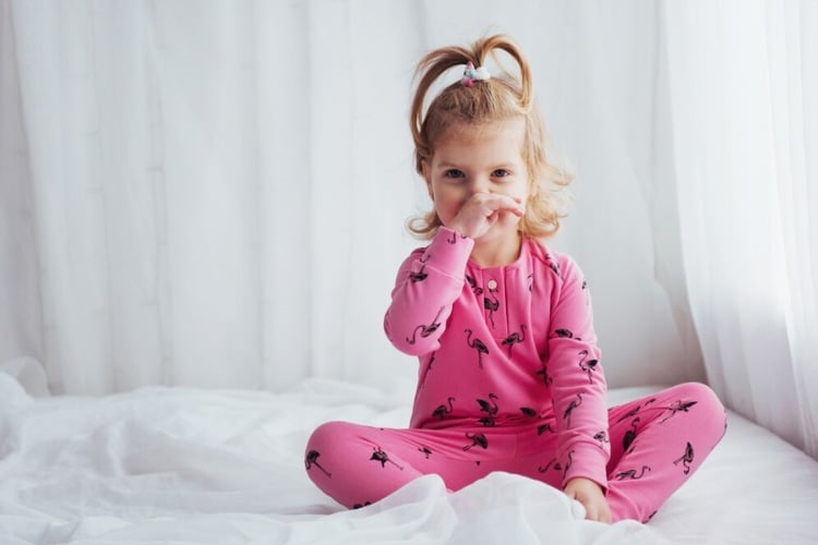 Menina de pele clara usando pijama rosa de calça e manga comprida, sentada sobre cama com lençóis brancos