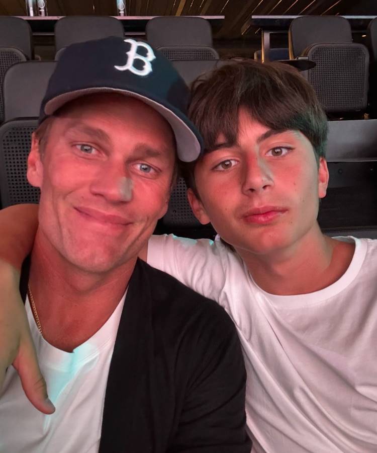 Tom Brady e Benjamin estão em um local fechado. O pai à esquerda usa boné preto com a letra "B" e camiseta branca, sorrindo levemente. O filho à direita veste camiseta branca e tem expressão neutra. Eles estão próximos, com o filho envolvendo o pai com o braço.