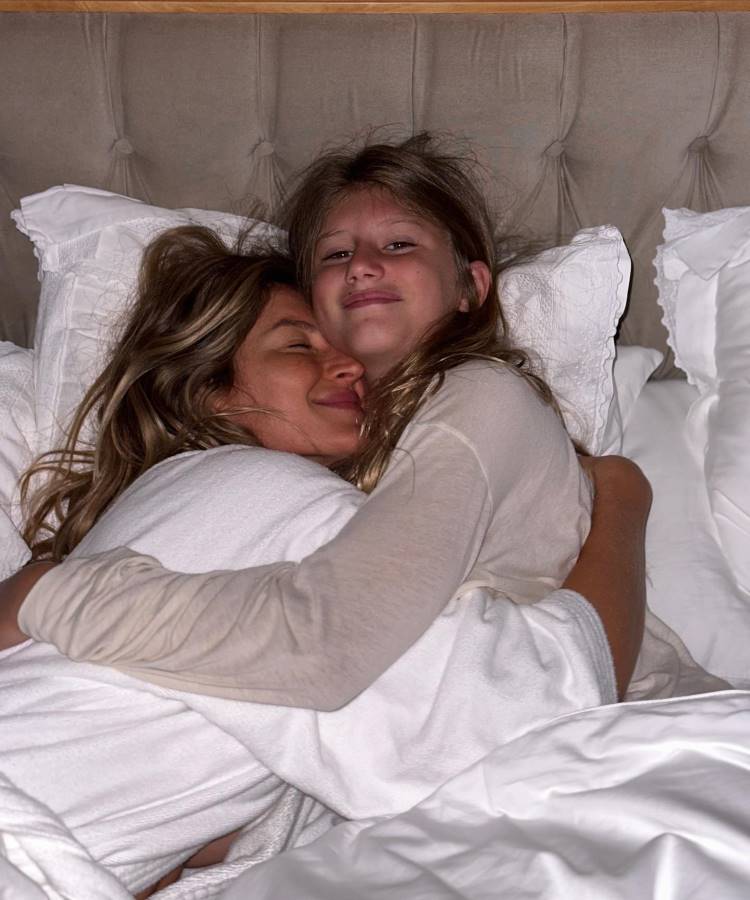 Gisele e Vivian estão deitadas em uma cama, abraçadas. A mulher, de olhos fechados, parece tranquila e feliz. A menina sorri suavemente, olhando para frente. Ambas estão envoltas em cobertores brancos, em um ambiente acolhedor e sereno, transmitindo amor e conforto.