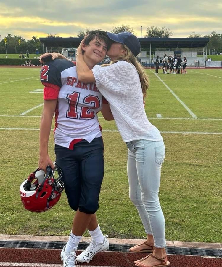 Um jovem jogador de futebol americano (Benjamin) sorri enquanto segura o capacete. Ele veste uniforme vermelho e branco com o número 12. Ao seu lado, uma mulher de calça jeans e blusa branca (Gisele Bündchen) o abraça e beija seu rosto. Estão em um campo de futebol, com pessoas ao fundo.