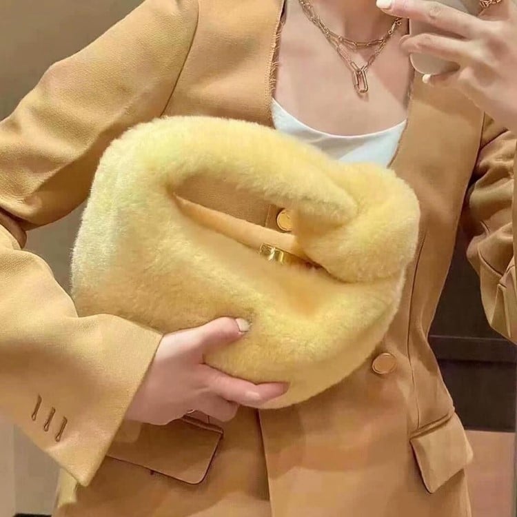 Uma mulher segura uma bolsa amarela felpuda, que complementa seu blazer da mesma cor. O design volumoso e a textura macia destacam-se como tendências em bolsas para 2025, que valorizam materiais táteis e formas ousadas. A combinação de tons quentes cria um visual acolhedor e moderno.