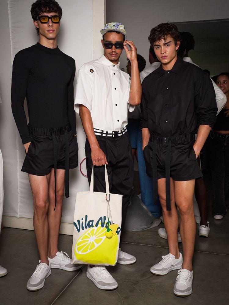 Foto de três homens. O da esquerda usa blusa de manga longa e bermuda, ambos pretos. O do meio usa camisa branca, calça preta e ecobag. O da direita usa camisa manga curta e bermuda, ambas pretas